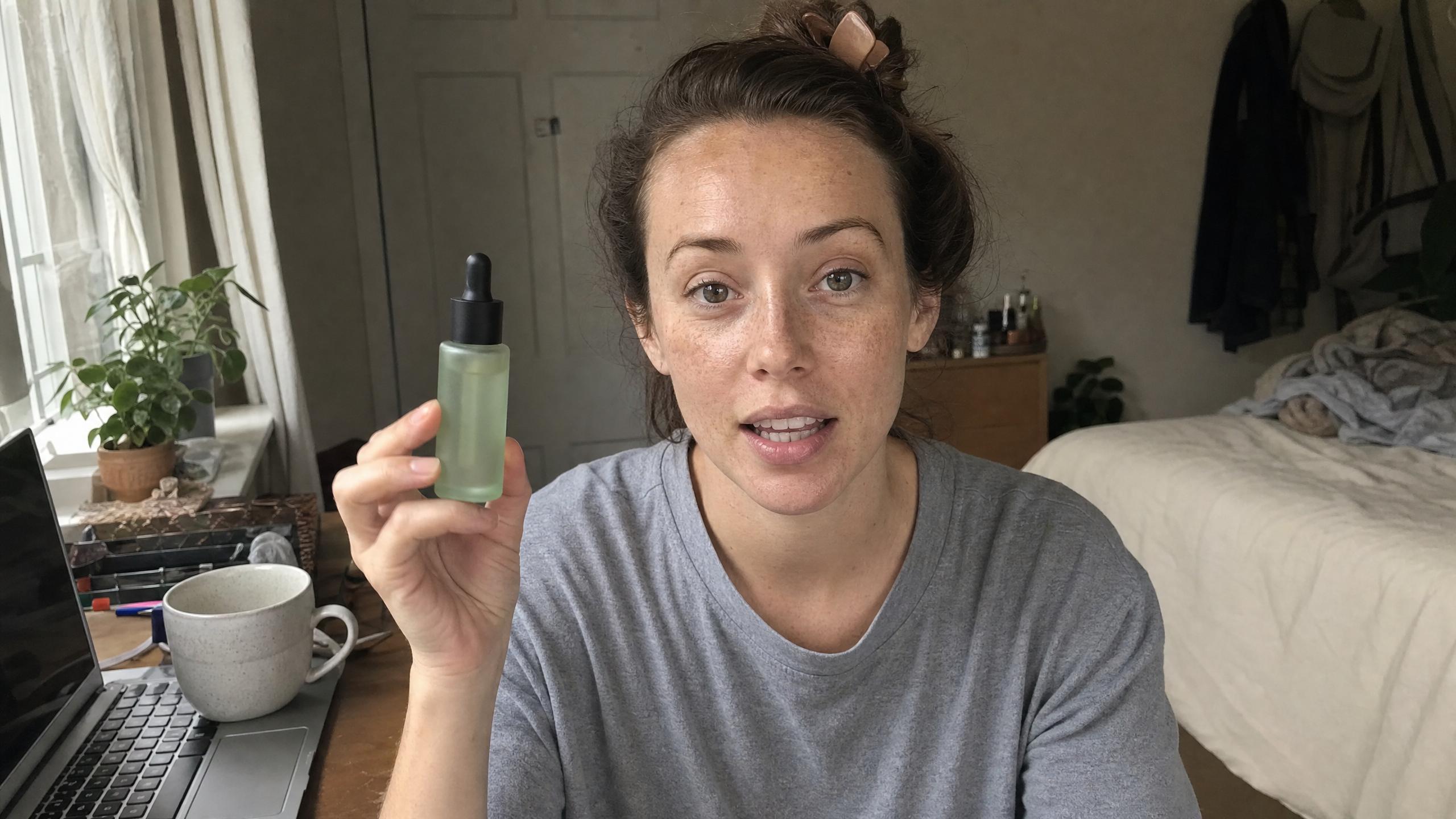 A freckled woman in a gray T-shirt holding up a green serum bottle at her desk in a bedroom