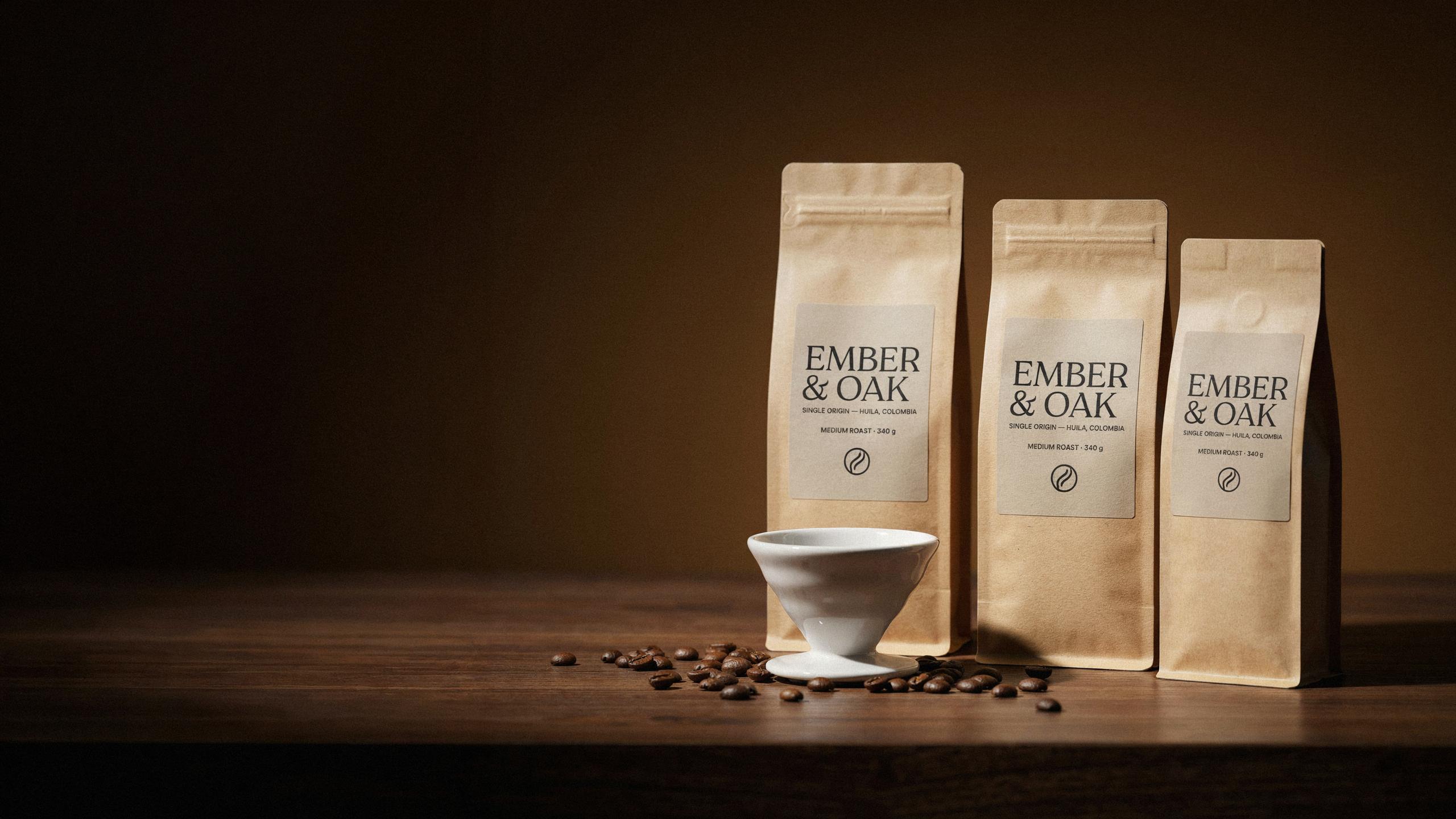 Three kraft-paper coffee bags labeled EMBER & OAK, Single Origin, Huila, Colombia, Medium Roast 340 g, stand on a walnut table beside a white ceramic pour-over dripper and scattered coffee beans.