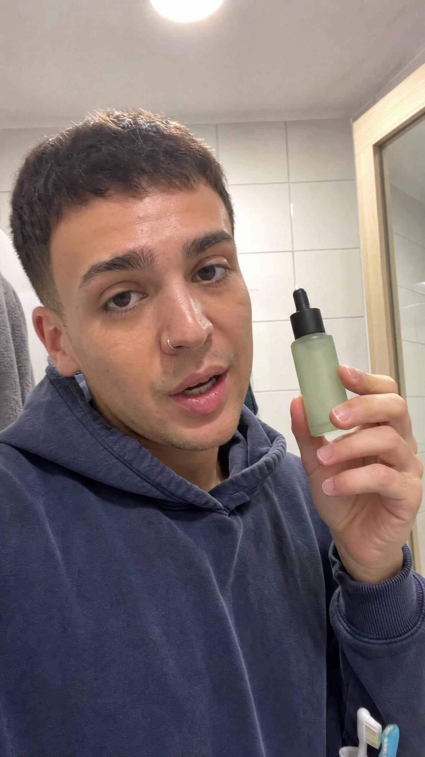 A young man in a navy hoodie holding a green dropper bottle while filming a selfie video in a bathroom