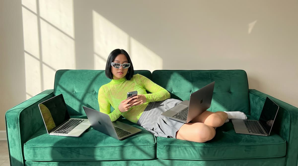 Person in a neon green top lounging on a green couch with a phone and four open laptops