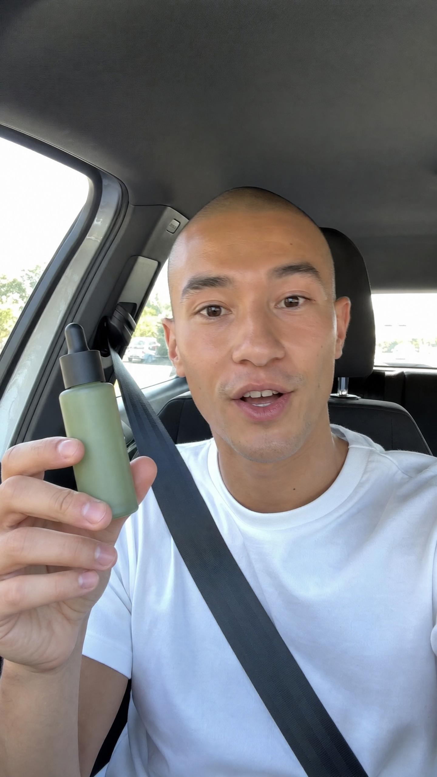 A man in a white T-shirt holding up a green dropper bottle while recording a selfie video in his car