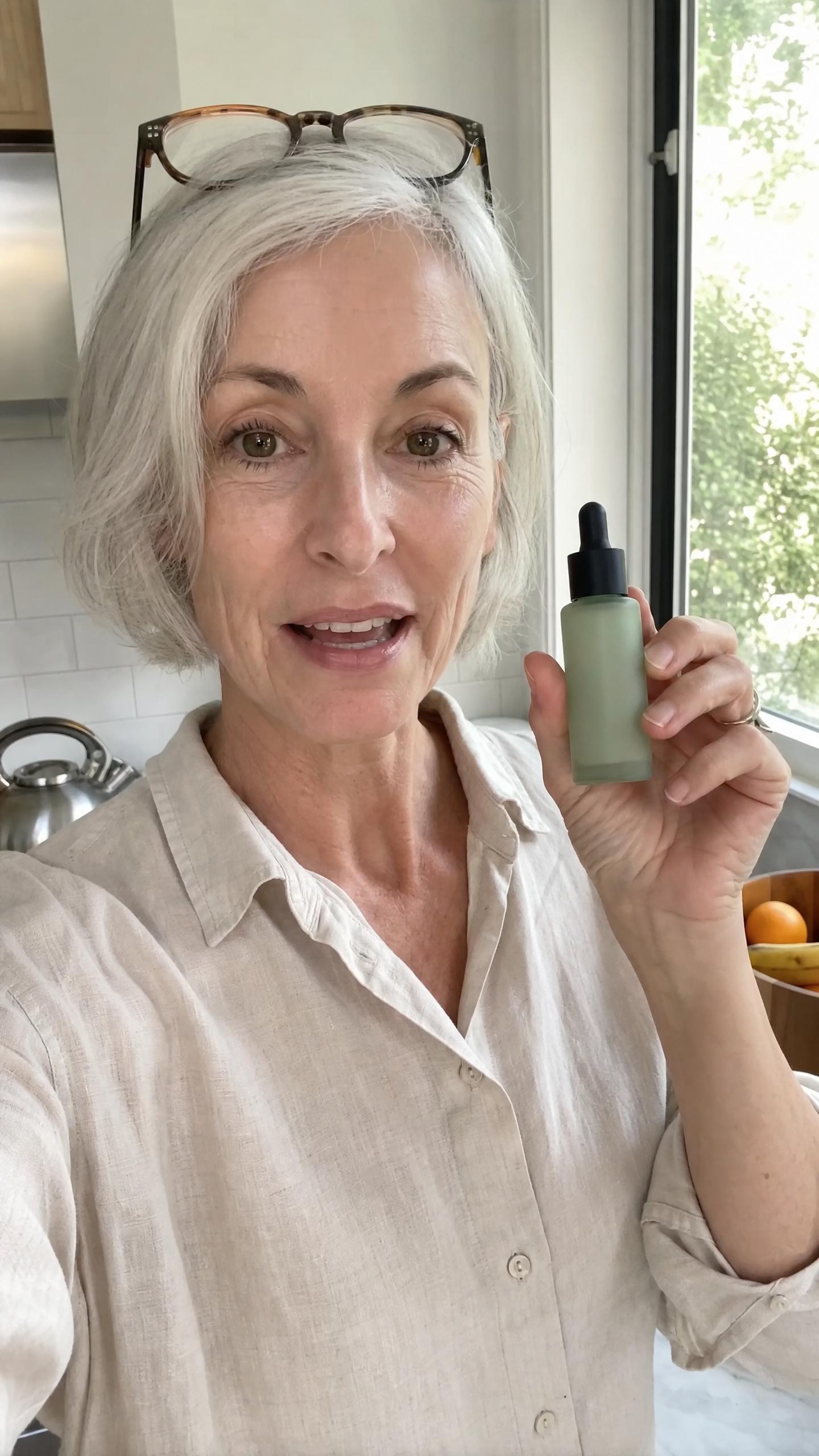 A silver-haired woman in a linen shirt holding up a green dropper bottle in a selfie video in her kitchen