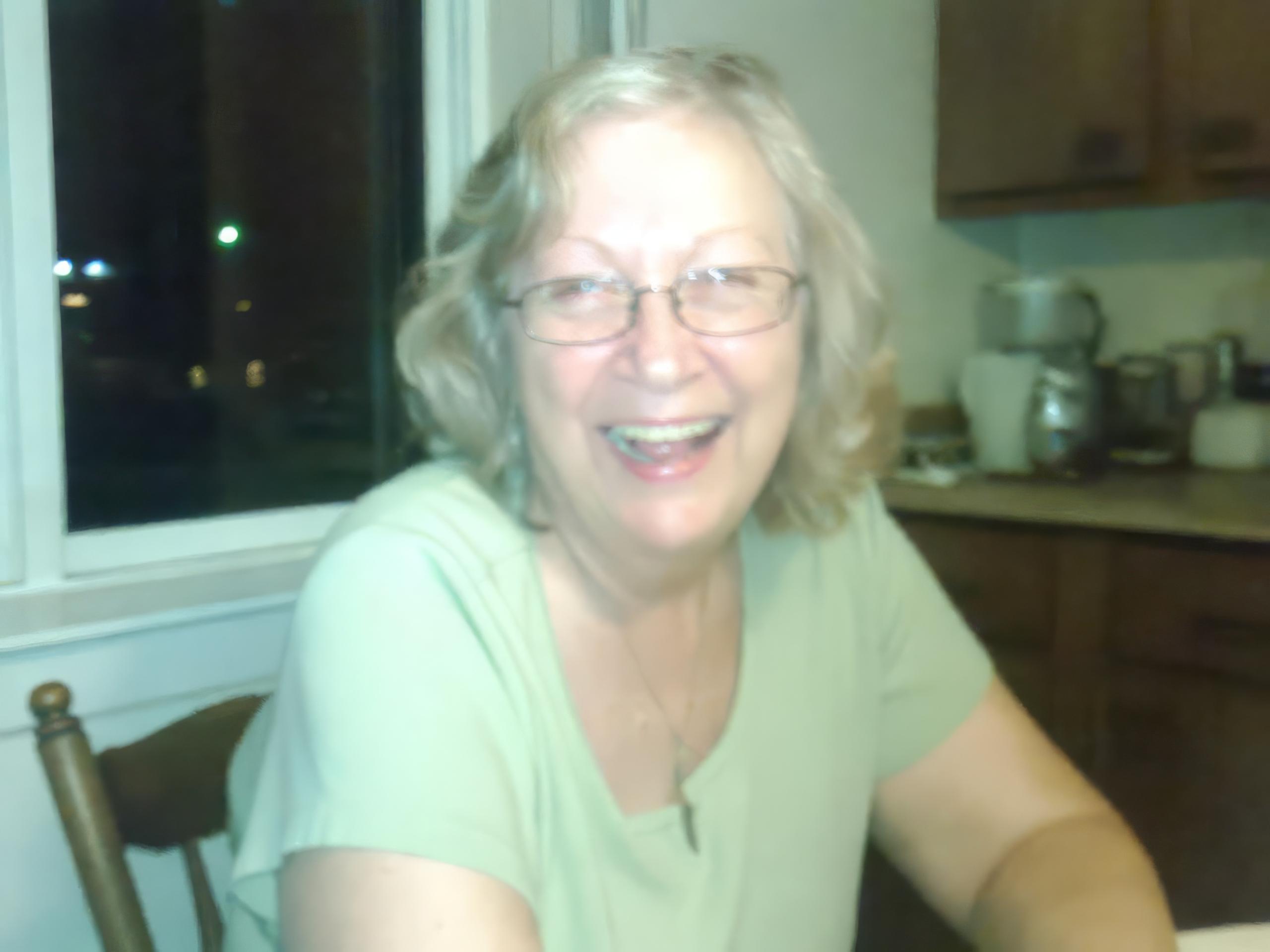 Slightly soft photo of a smiling gray-haired woman in glasses and a green top in a kitchen