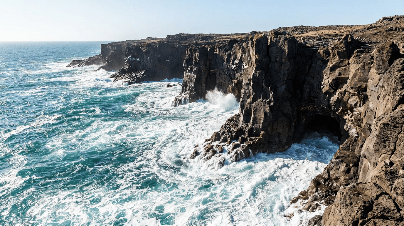 Waves foam against dark rocky sea cliffs under a clear sky, with a cave opening at the base of the cliff