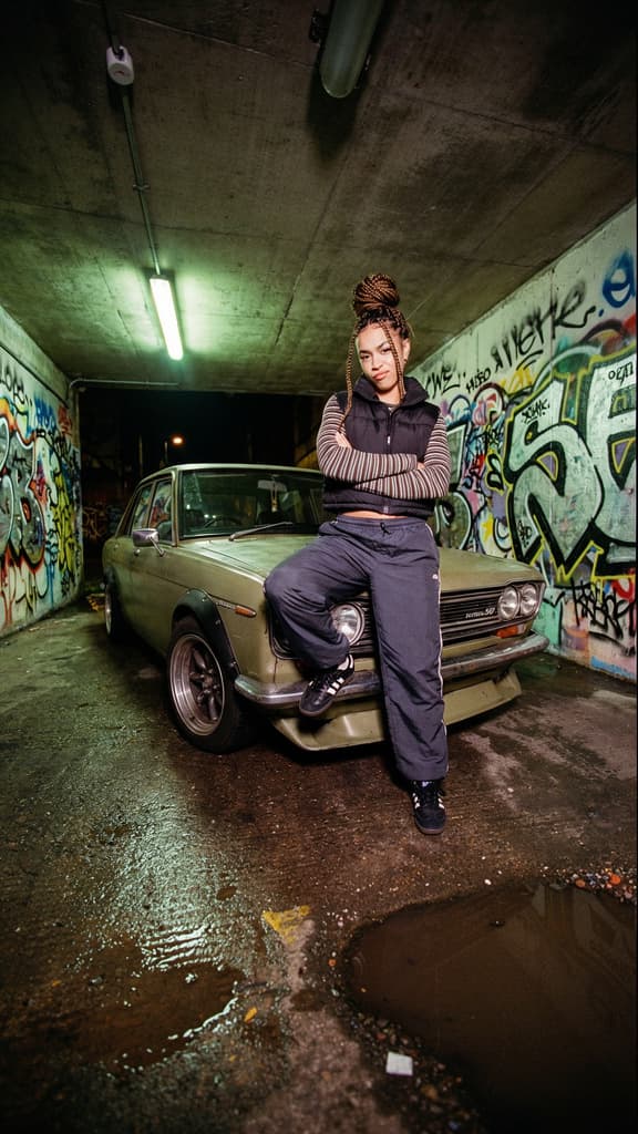 A full-length vertical image of a woman with braided hair in a high bun leaning on a vintage olive-green sedan. She wears a black puffer vest and track pants. The setting is a concrete tunnel decorated with graffiti, featuring wet ground with a puddle, generated using Flux.2 [pro] at 576x1024 resolution.