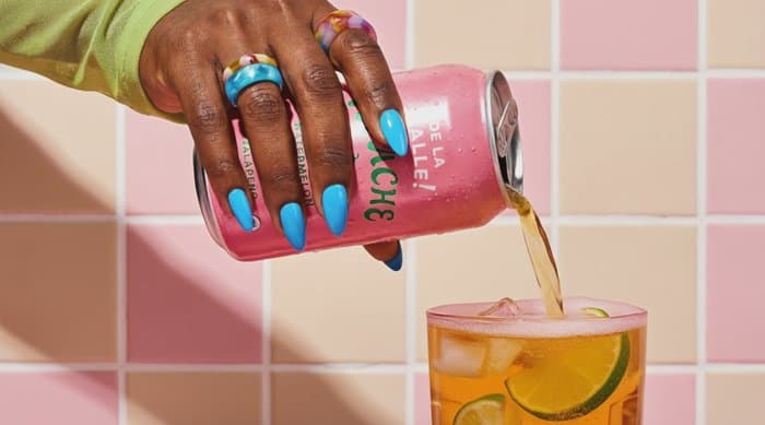 A hand pouring a canned drink over ice against a pink tiled wall