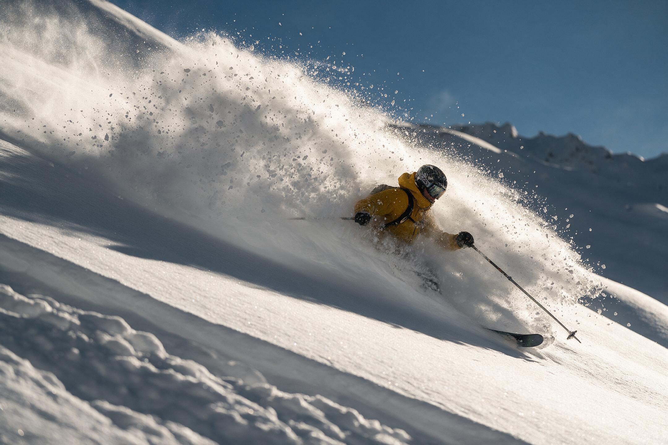 Skier in a mustard jacket carving through deep powder against a cobalt sky