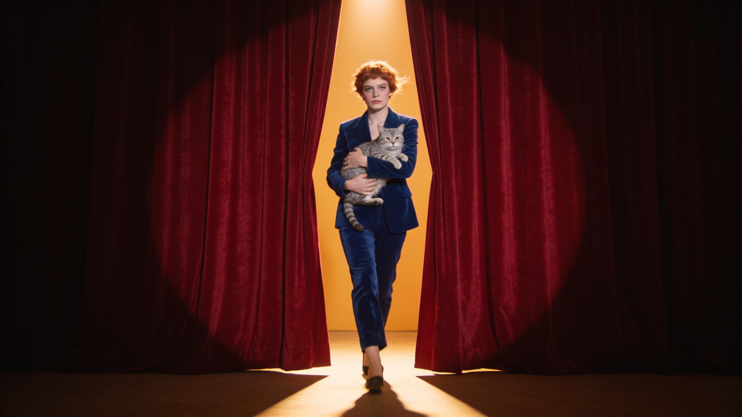 The woman in the cobalt velvet suit walking through parted red stage curtains carrying a tabby cat