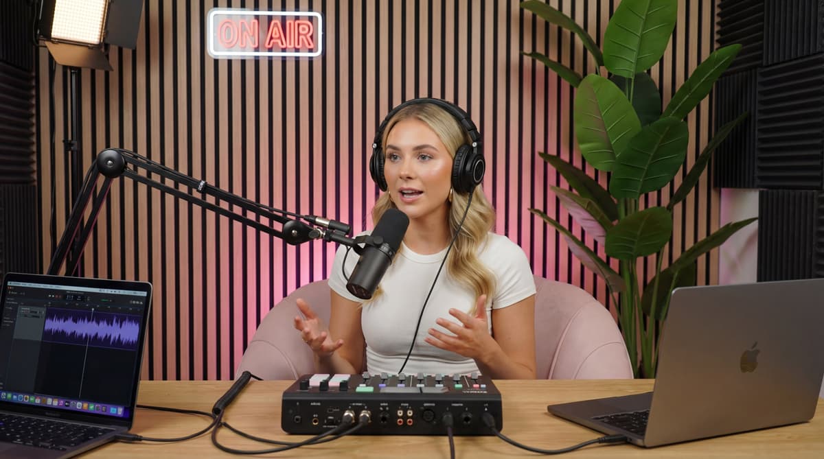 Woman wearing headphones speaks into a boom microphone at a podcast desk with mixer, laptops, and "On Air" sign