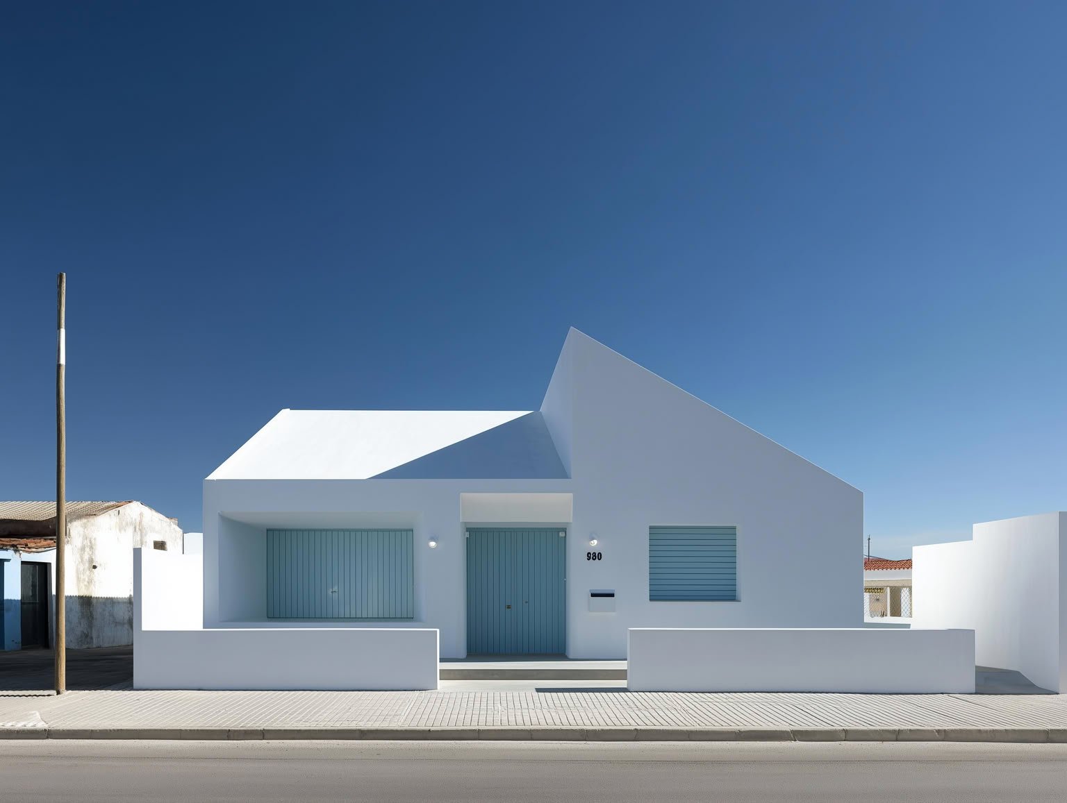 White minimalist house with blue doors and shutters, featuring an asymmetrical roof, under a clear blue sky.