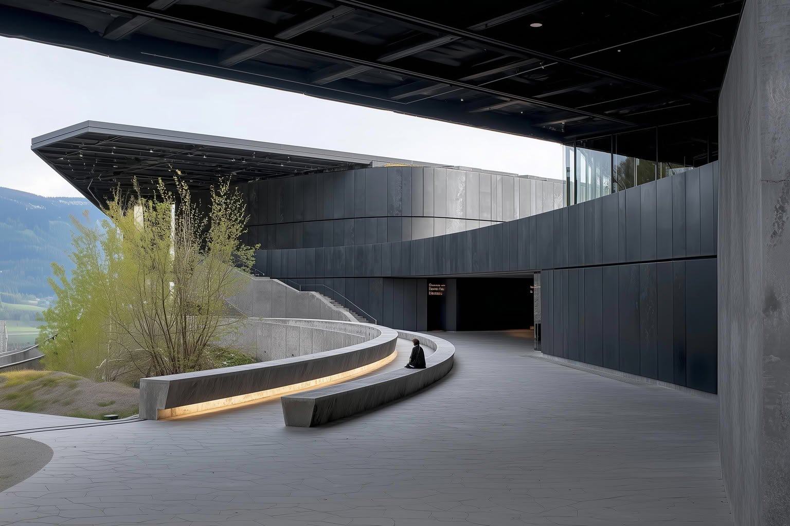 Modern building with dark gray panels, curved concrete path, integrated lighting, and a person sitting on a bench. Mountains.