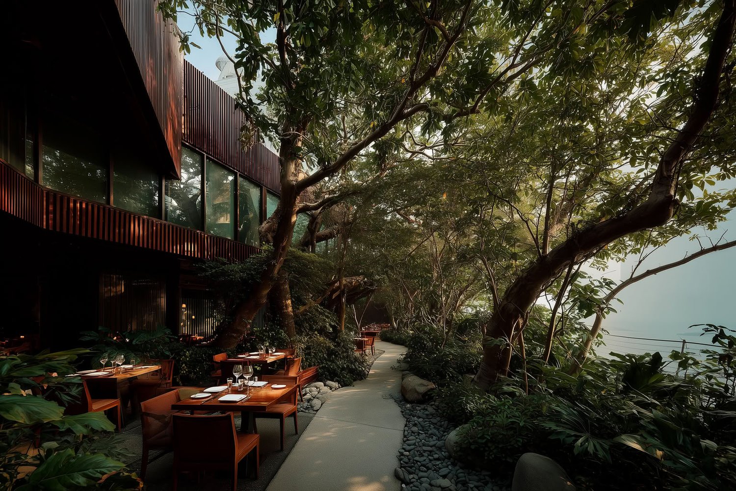 Outdoor restaurant with dark wood architecture, lush greenery, a stone pathway, and dining tables by the water.