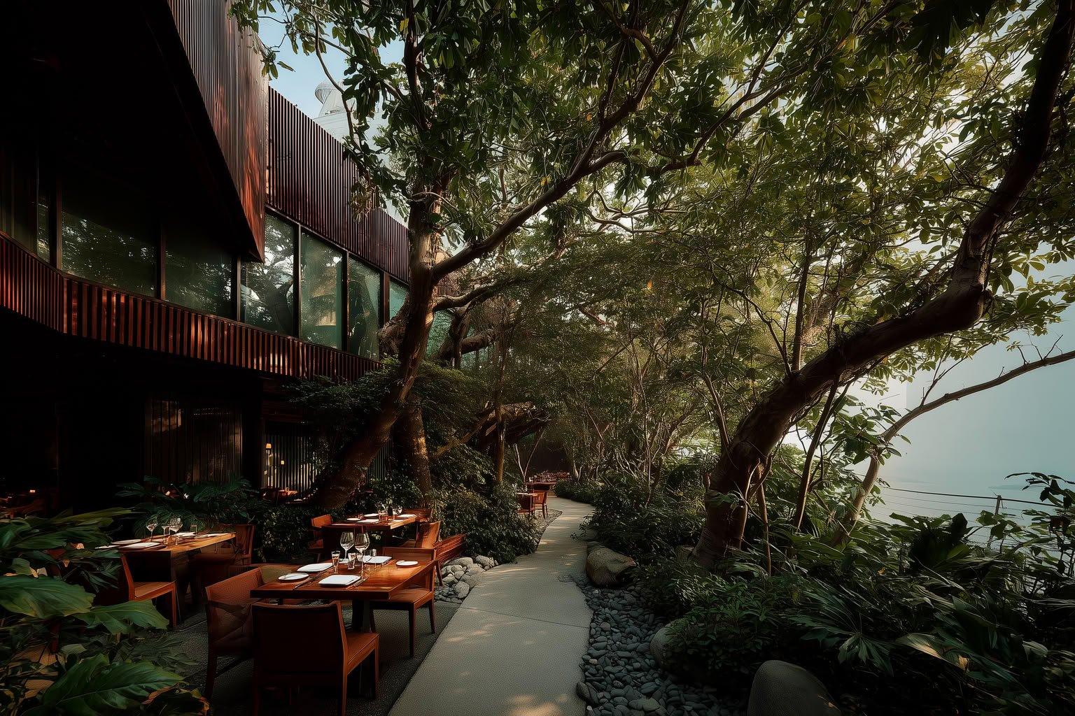Outdoor restaurant with dark wood architecture, lush greenery, a stone pathway, and dining tables by the water.