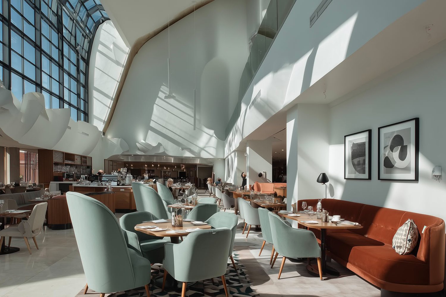 Spacious, modern restaurant interior with large curved glass windows, mint green chairs, and a rust-colored banquette, bathed in natural light.
