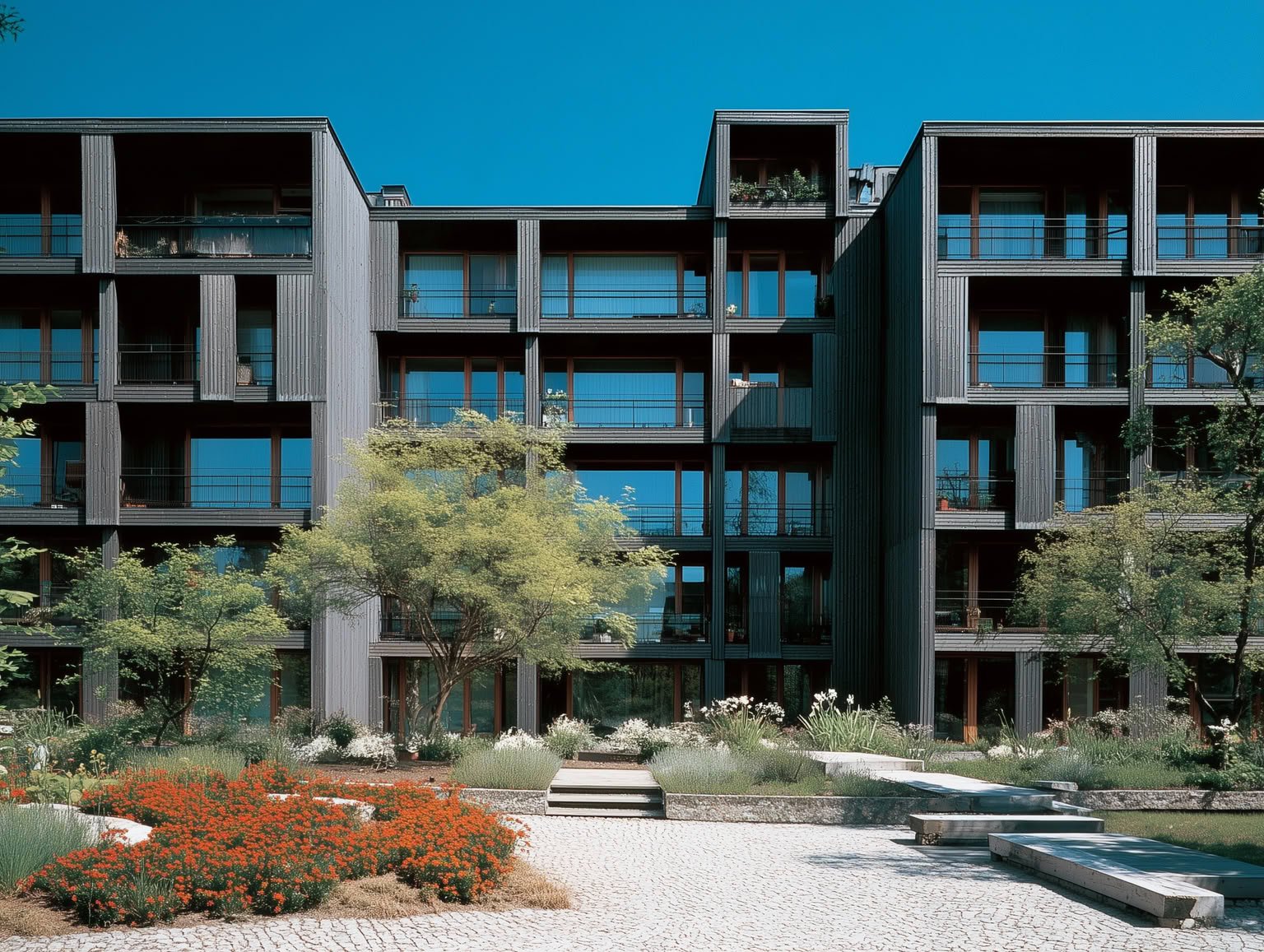 Multi-story dark timber-clad buildings with large windows, balconies, and an extensive landscaped garden featuring red flowers and trees.