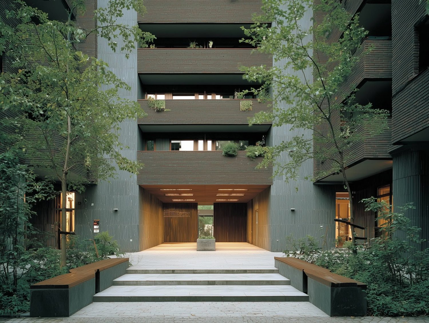 Modern building entrance with textured dark facade, grey panels, warm wood, and green trees.
