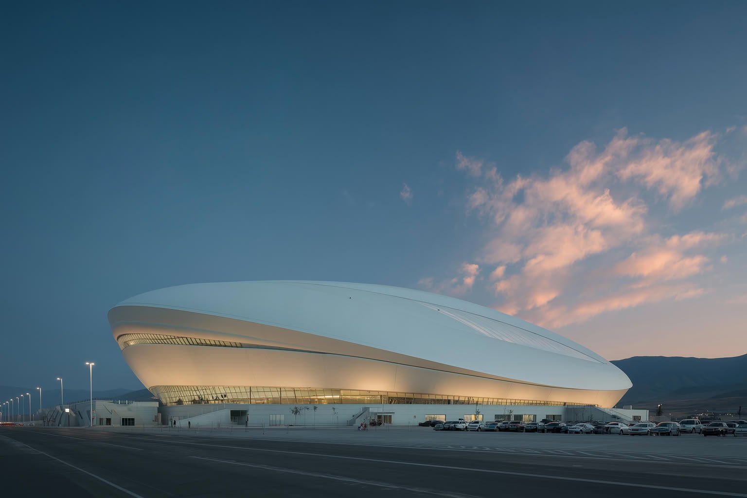 Modern white elliptical building with illuminated glass base at dusk under a blue and pink sky with clouds and distant mountains.