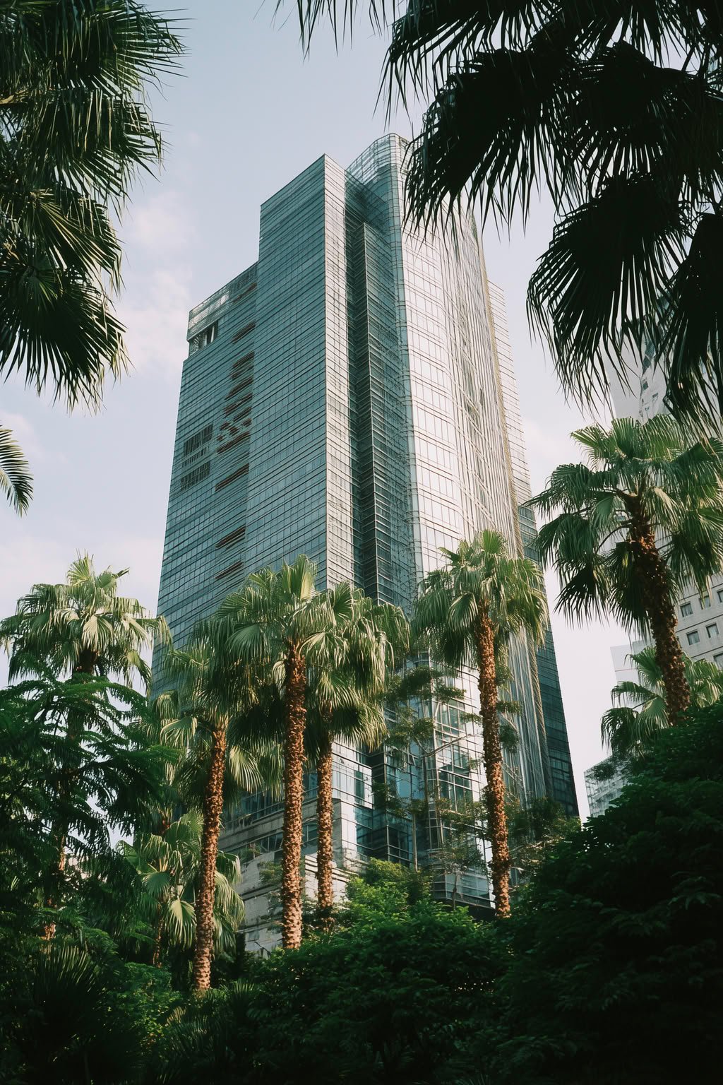 Modern glass skyscraper rises amidst lush green palm trees under a bright sky.