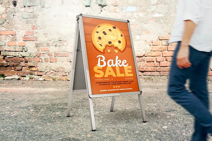 A passerby walks by a bright orange Bake Sale sign featuring a large cookie graphic, created using Poster Maker Machines.