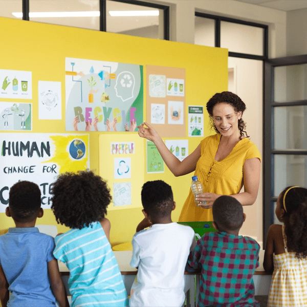 A teacher stands before colorful posters in a classroom, illustrating recycling and sustainability concepts to a group of young students.