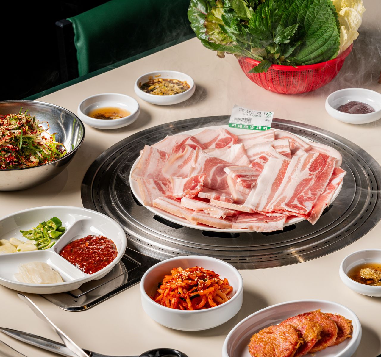 고기집 프랜차이즈 음식사진 촬영 - 원형 불판 위 얇게 썬 삼겹살 한 판과 쌈채소 바구니, 파채·무생채·고추와 마늘 찬을 차린 한 상