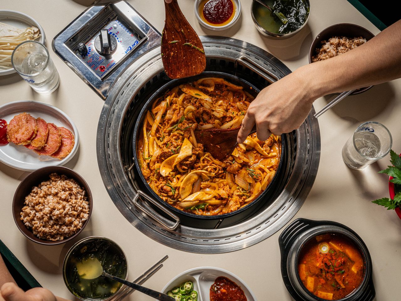 고기집 프랜차이즈 음식사진 촬영 - 바로 위에서 본 불판, 나무 주걱으로 볶음을 뒤적이는 손과 둘레의 잡곡밥·미역국·찌개