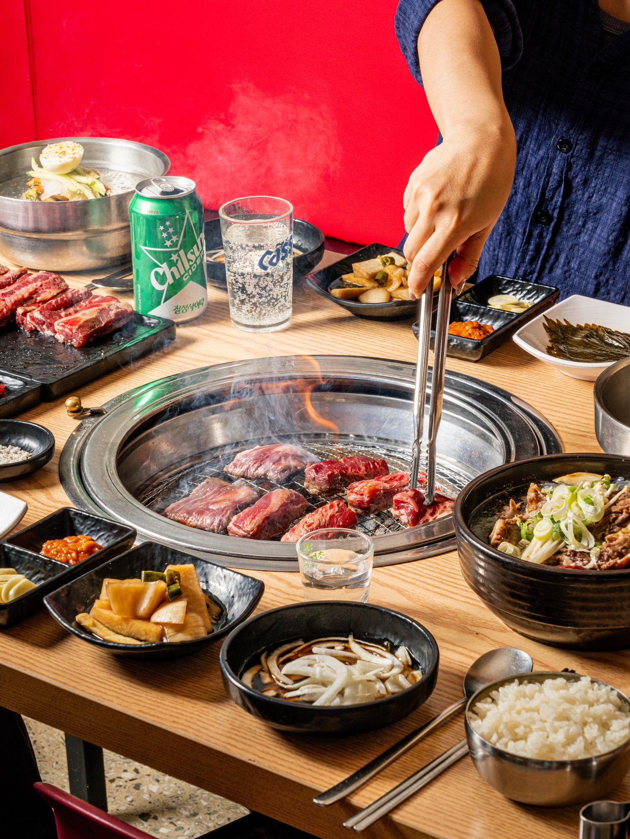 고기집 연출 촬영 - 붉은 벽 앞 숯불 석쇠에서 집게로 고기를 뒤집는 장면과 장아찌, 공기밥이 놓인 테이블