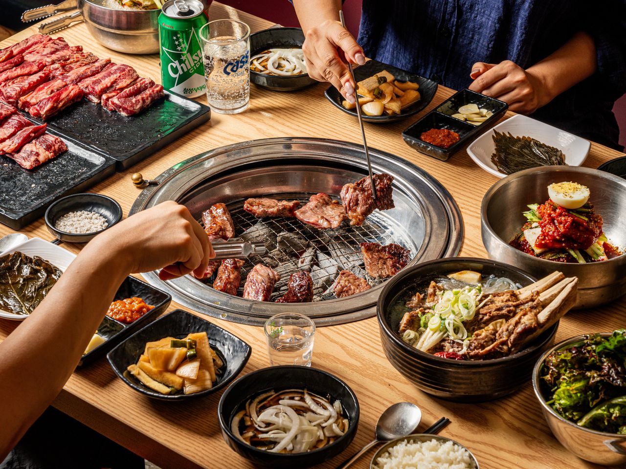 고기집 연출 촬영 - 두 사람이 집게로 고기를 굽고 원육 보드, 비빔냉면, 갈비탕, 쌈채소가 둘러싼 한상