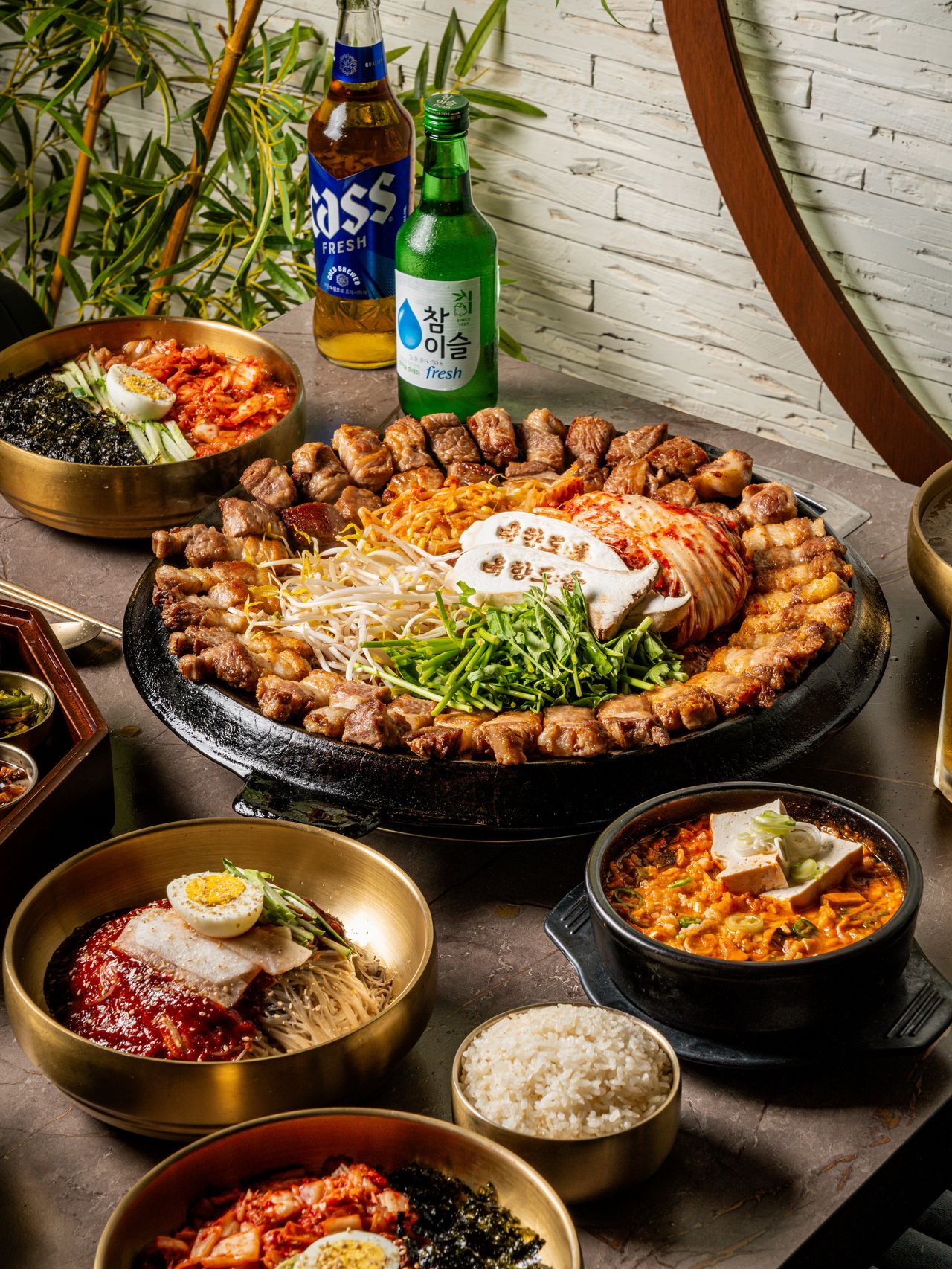 고기집 출장 음식사진 촬영 - 흰 벽돌 벽과 대나무 화분 앞, 맥주·소주 두 병 뒤로 불판 한 상과 냉면·찌개·공기밥을 차린 세로 컷