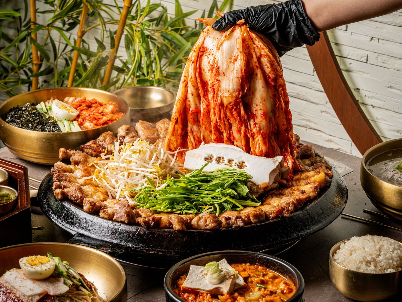 고기집 출장 음식사진 촬영 - 장갑 낀 손이 통김치를 들어 올리고 불판 위에 미나리·콩나물이 놓인 컷