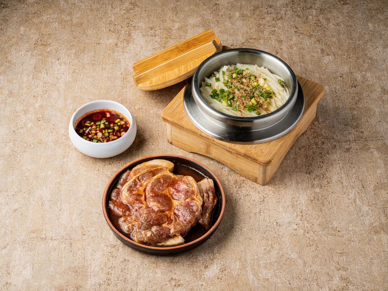 산솔 신사본점 메뉴 촬영 - 베이지 스톤 배경 위 양념 목살 접시와 나무 받침에 올린 채 무침 그릇, 간장 소스