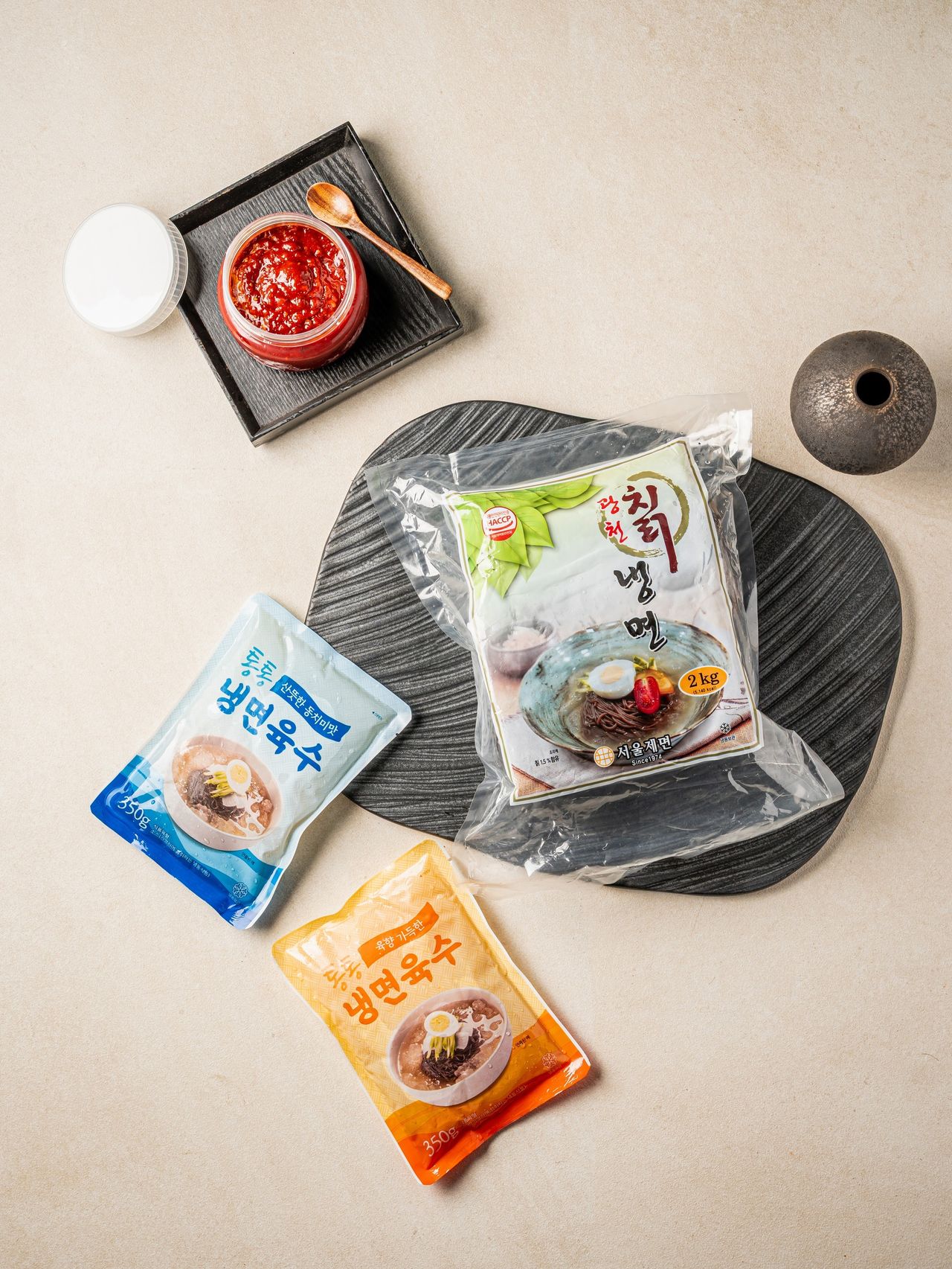 스튜디오 냉면 상세페이지 촬영 - 진공 포장 면 봉지와 두 가지 색 육수 파우치, 양념장 병을 짙은 돌판 위에 놓은 패키지 플랫레이