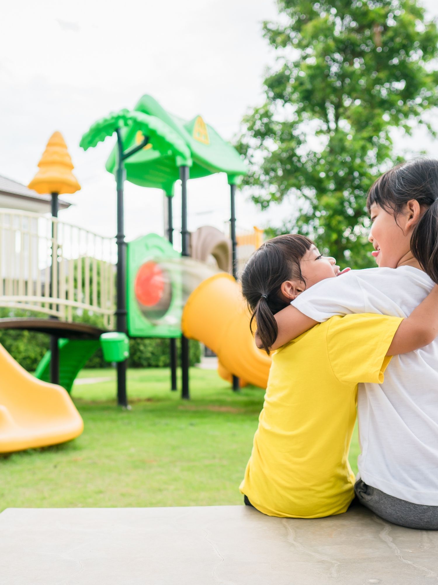 Two young children hug on a bench beside a playground.
