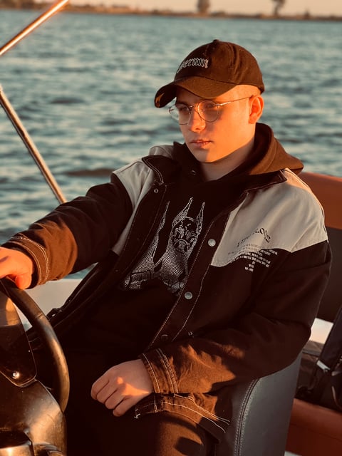 Man with regular cap, black hoodie and black jeans jacket driving a boat on the lake during sunset