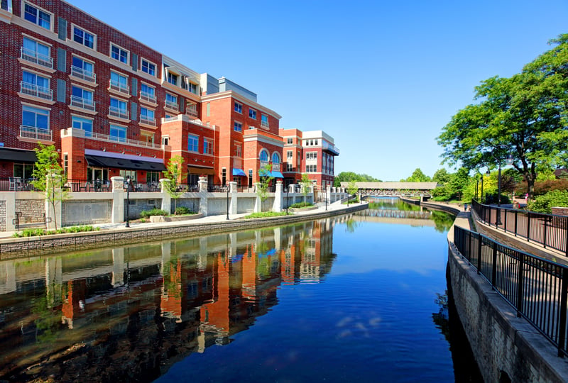 Downtown Naperville, Illinois, with riverfront buildings, trees and a pedestrian walkway.
