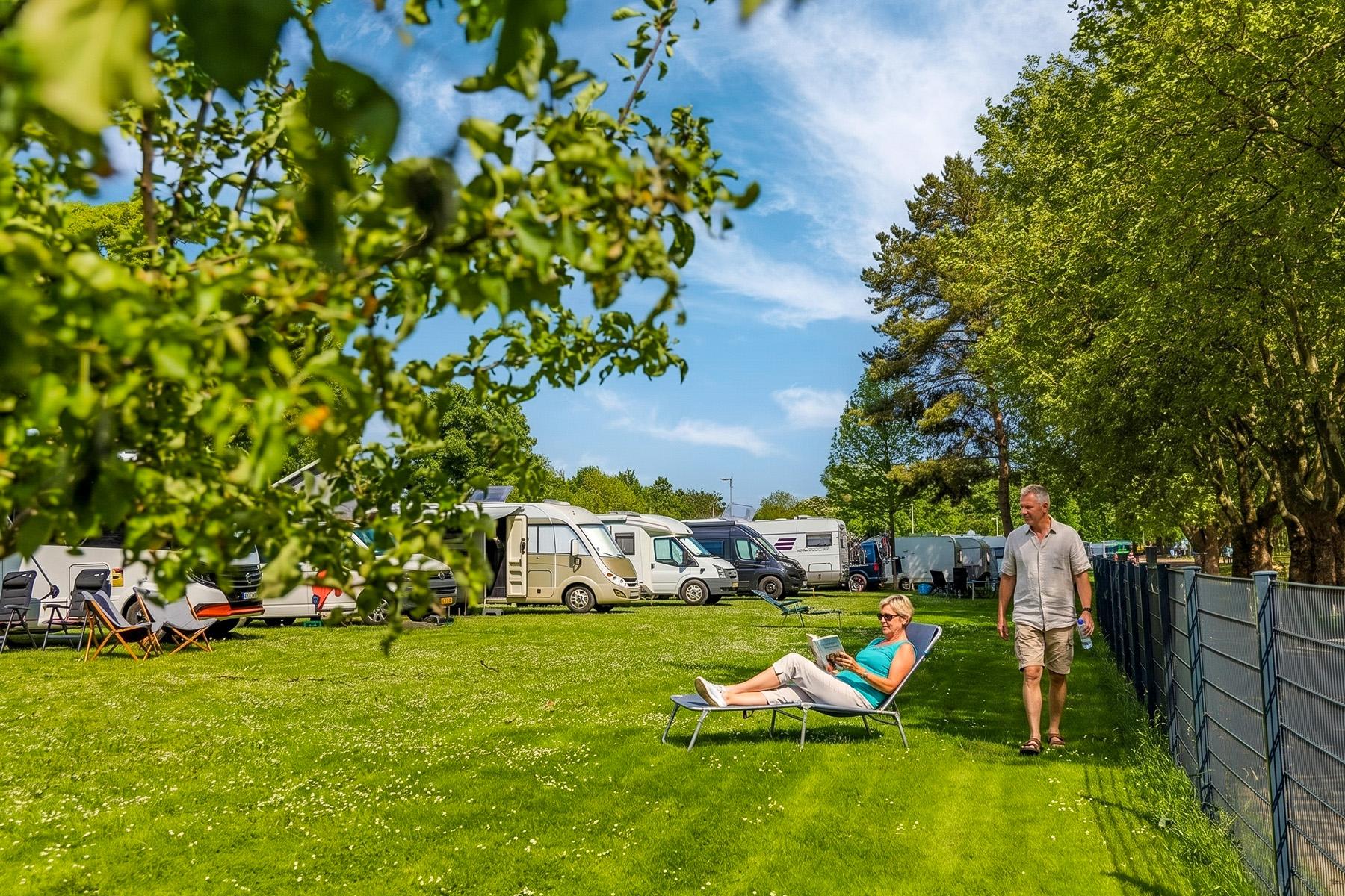 Rheingaucamping Geisenheim – kvinde slapper af på liggestol i skyggen af træer, mand står ved siden af, pladser med campingvogne og autocampere i baggrunden