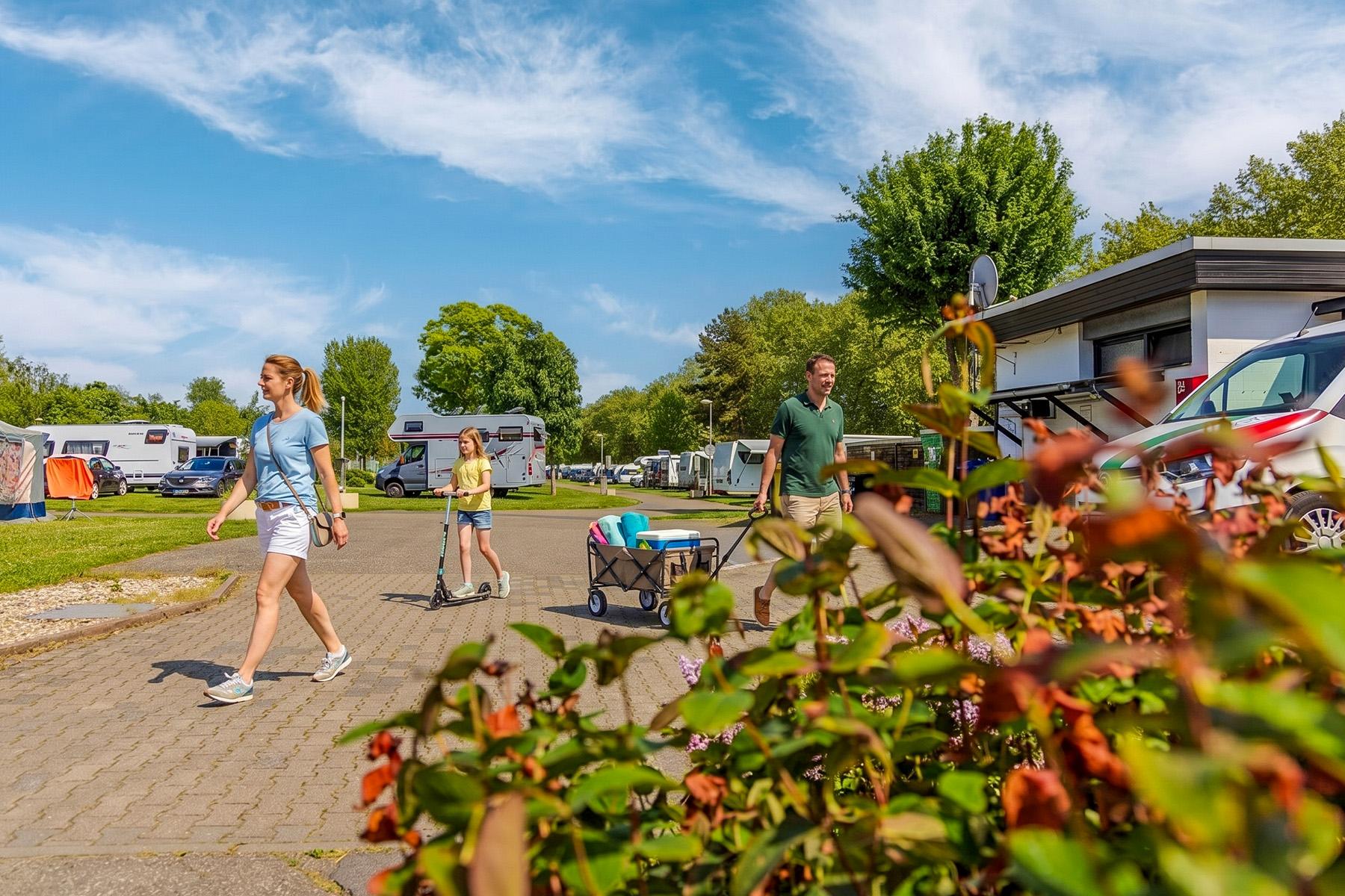 Rheingaucamping Geisenheim – familie på campingstien: mor, barn på løbehjul og far med håndvogn, campingvogne og autocampere på pladser i baggrunden