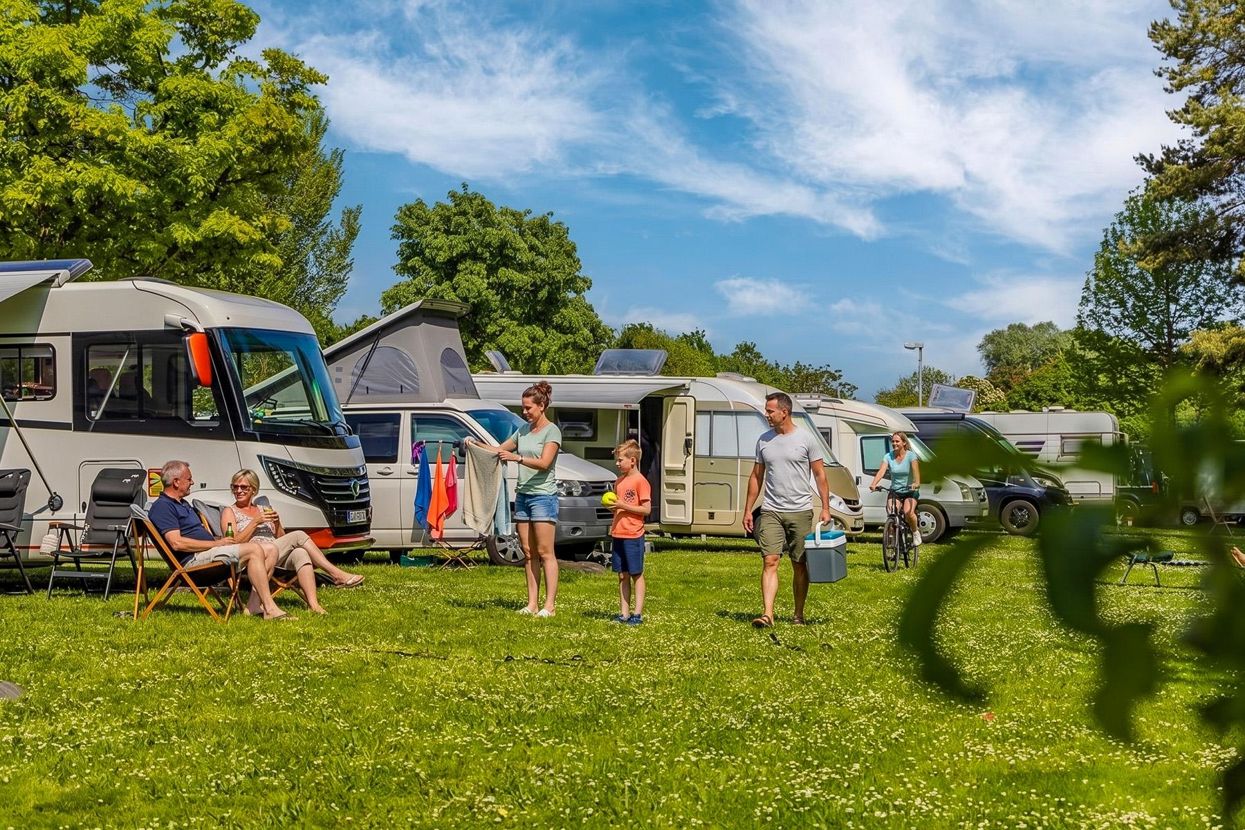 Rheingaucamping Geisenheim – livlig campingatmosfære: par i campingstole, legende barn med mor og mand med køleboks foran flere autocampere på grønt græs