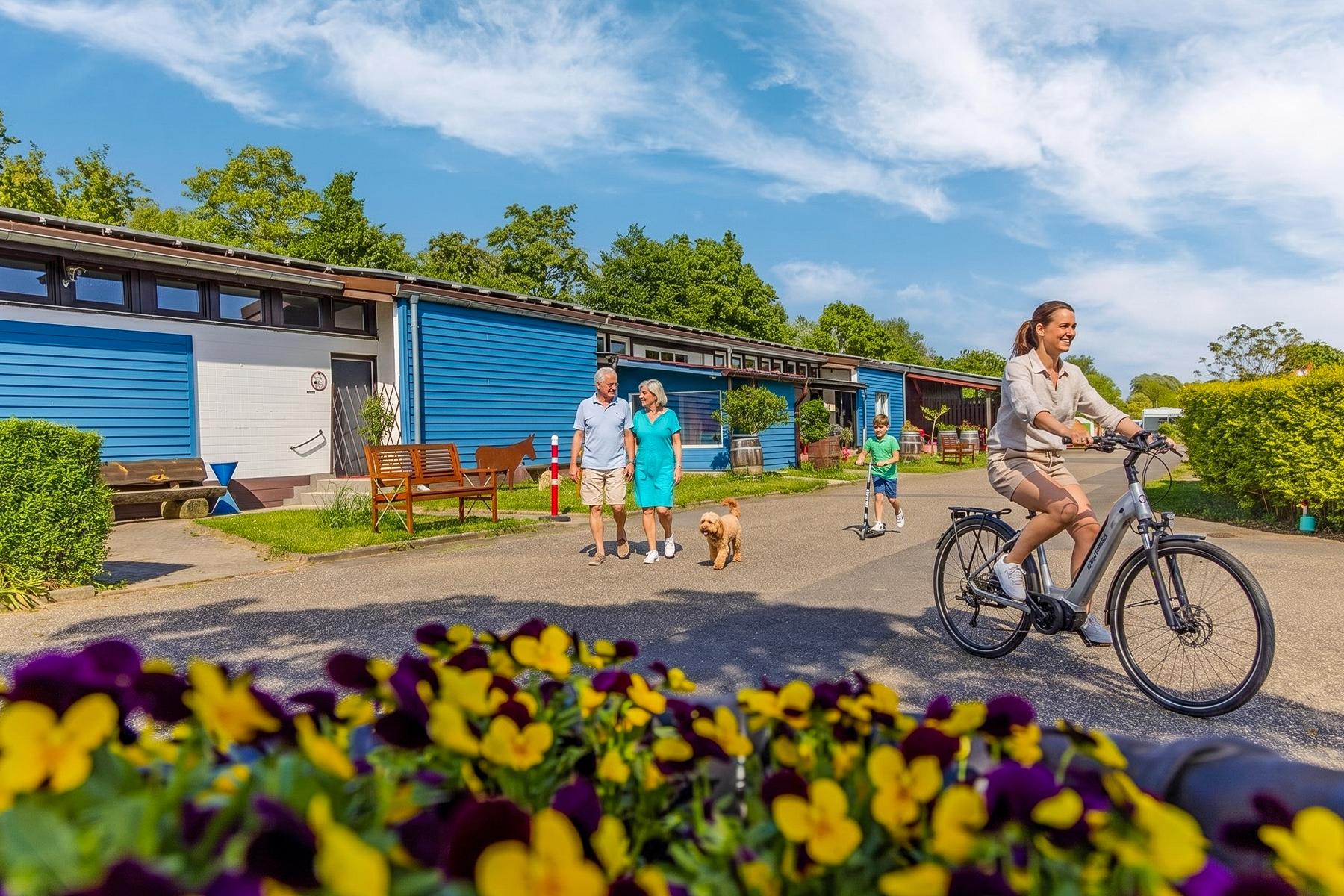 Rheingaucamping Geisenheim – feriegæst på el-cykel og gæstepar med hund foran sanitærbygningen, farverige stedmoderblomster i forgrunden