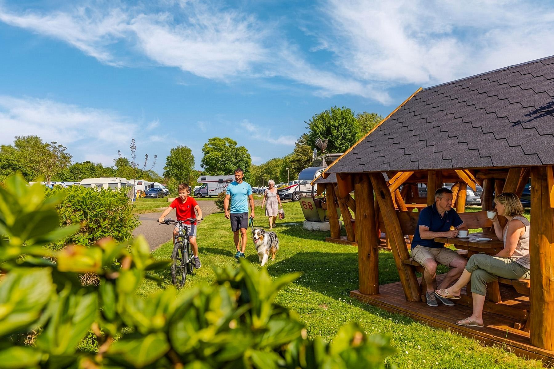 Rheingaucamping Geisenheim – par under træpavilion på campingpladsen, feriegæst på cykel og mand med hund på vej mellem pladserne i baggrunden
