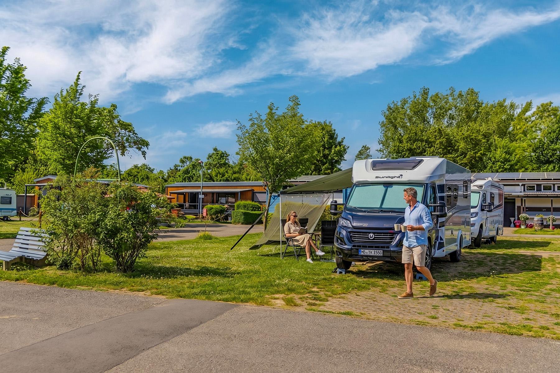 Rheingaucamping Geisenheim – par på autocamper-pladsen: mand står foran køretøjet, kvinde sidder under markisen i campingstol, sanitærbygning og træer i baggrunden