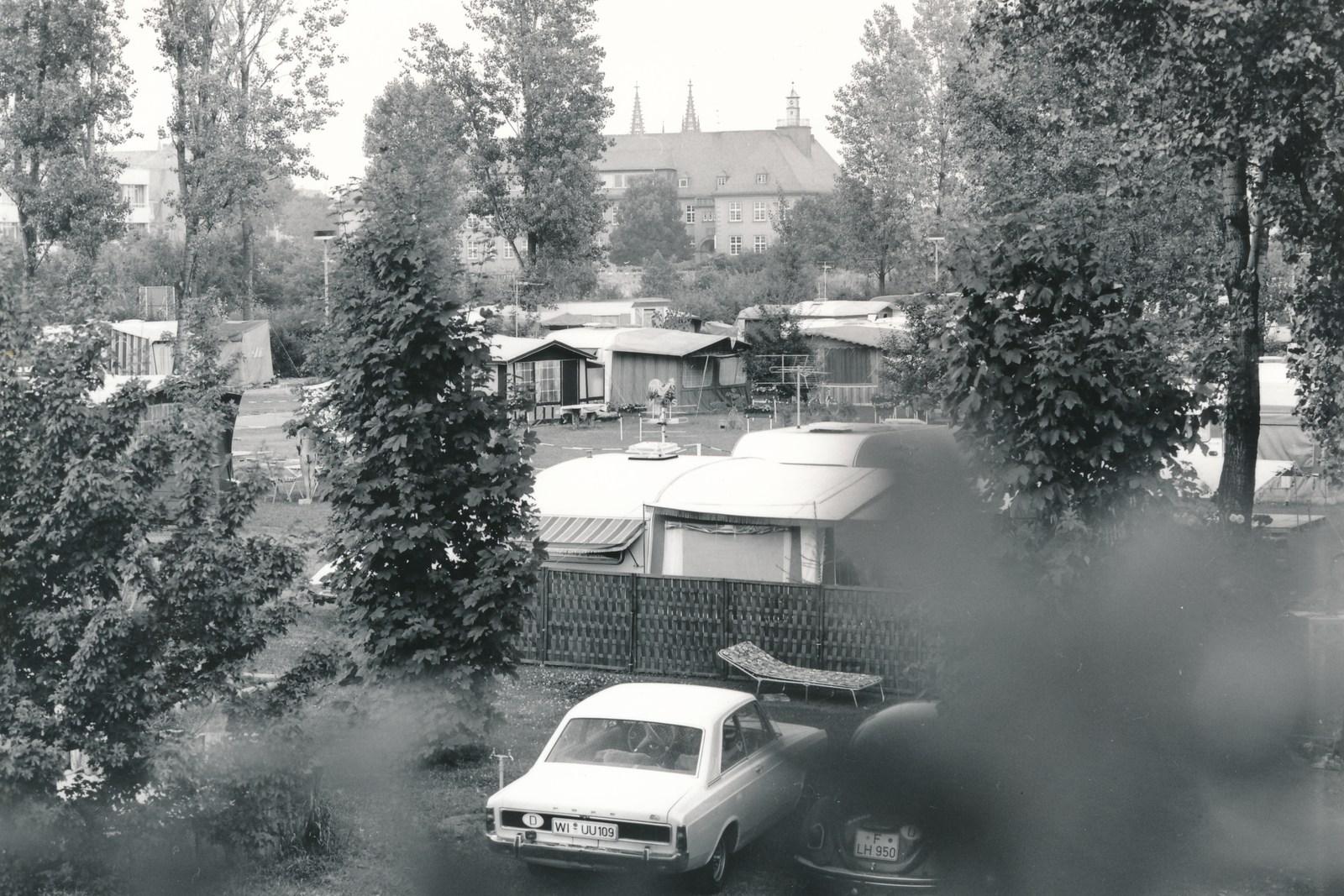 Caravans en een auto uit de jaren zeventig op de camping, daarachter bomen en een kerktoren