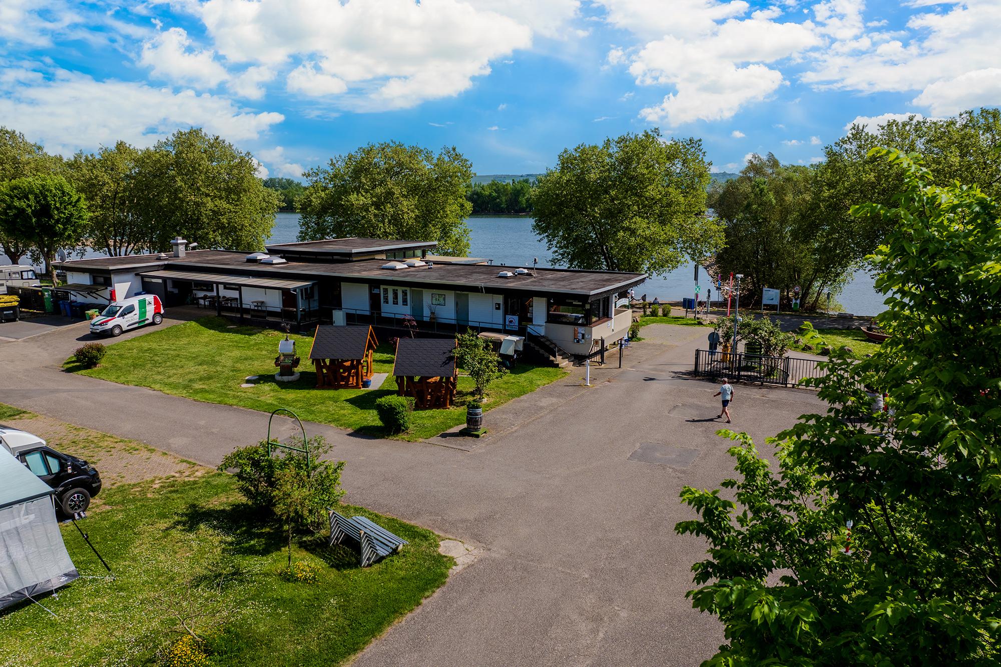 Luftfoto av innkjørselen og resepsjonsområdet på Rheingaucamping Geisenheim med sanitærbygning, parkeringsplass og utsikt til Rhinen