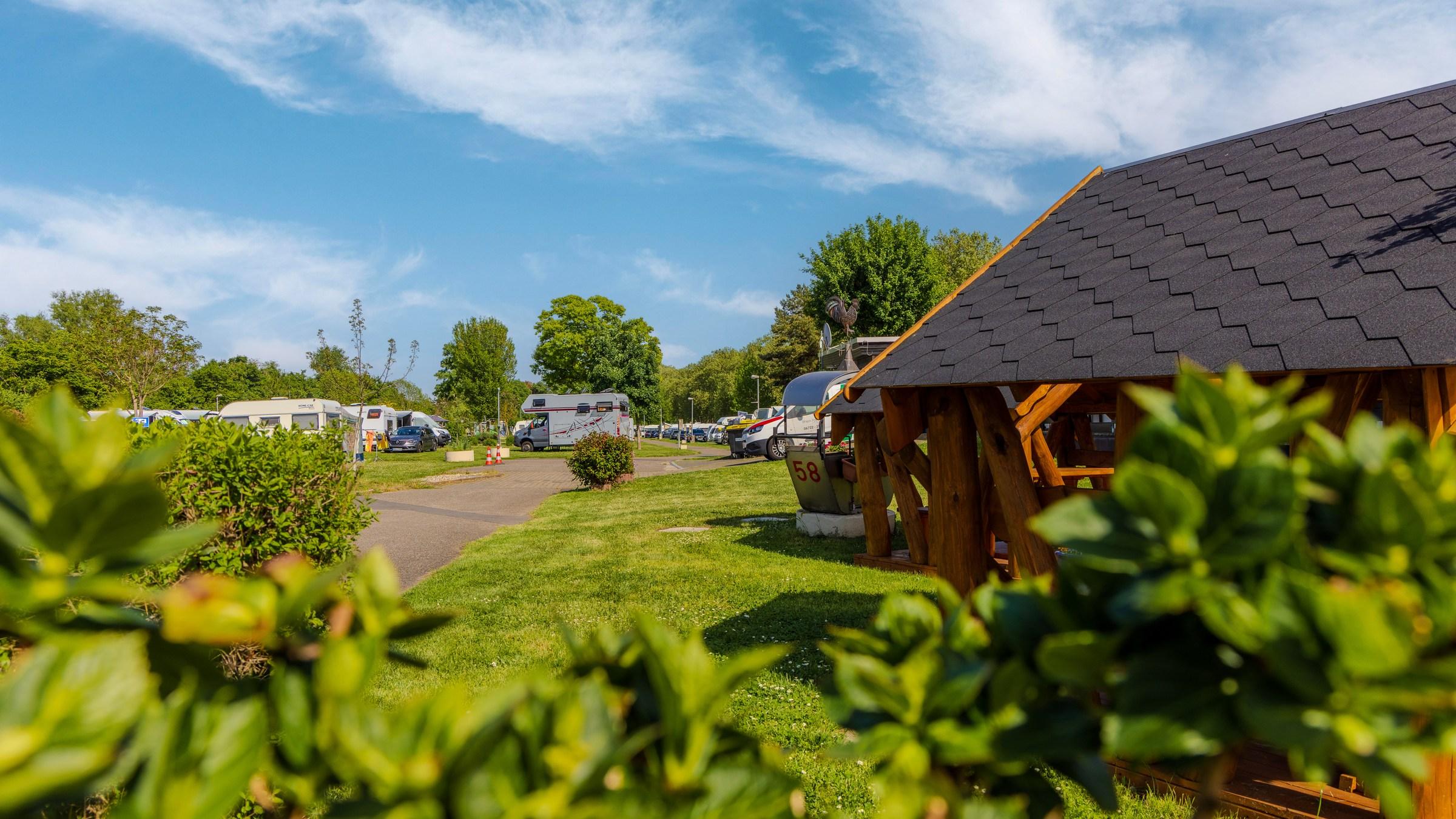 Rheingaucamping Geisenheim vandaag – over het gras naar het houten paviljoen, daarachter standplaatsen met campers