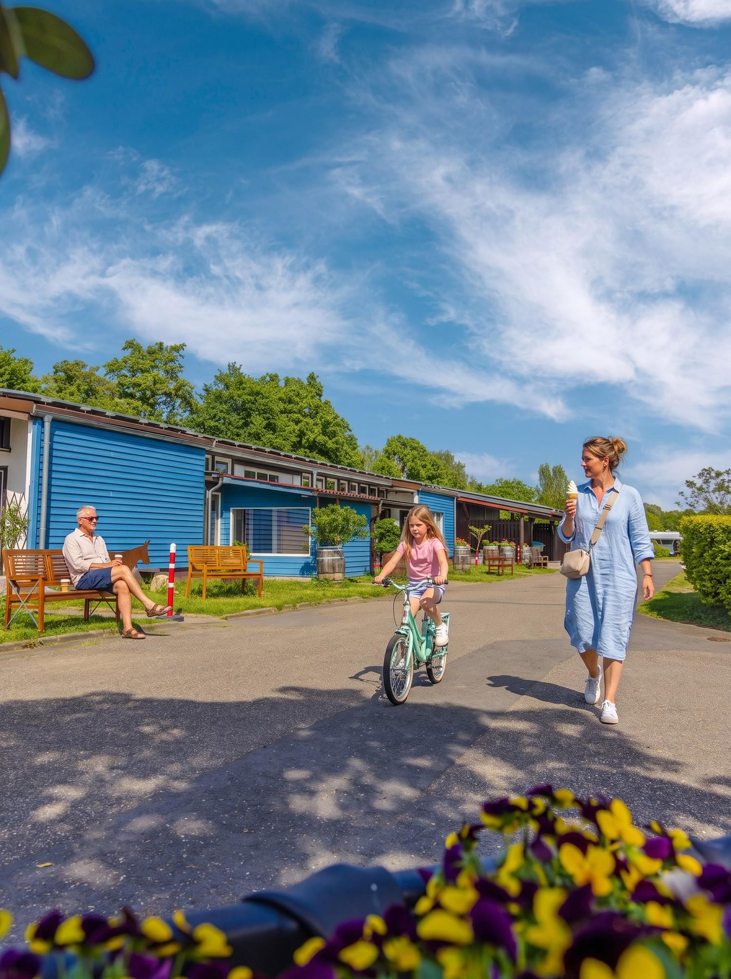 Rheingaucamping Geisenheim – vy över de blå sanitetsbyggnaderna, en flicka cyklar och en kvinna med glass går bredvid